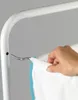A hand is shown attaching a white fabric bag with a blue trim to a silver hook on a white metal frame. Produktbild-Vorschau