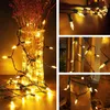 A collage of images showing warm white LED string lights in various settings, including wrapped around a vase with dried flowers, draped on wooden planks, and wrapped around a tree trunk. Produktbild-Vorschau
