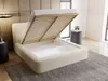 A beige upholstered bed with its storage compartment open, revealing the slatted base and storage space. The bed is in a bedroom setting with a rug and a bedside table. Produktbild-Vorschau