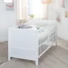A white baby crib with bedding and a small toy is shown in a nursery setting with artwork on the walls and a window with sheer curtains. Produktbild-Vorschau