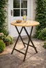 A round wooden table with a metal base is set up on a patio, with a bottle of water and two glasses of wine on it. Plants and a white door are in the background. Produktbild-Vorschau
