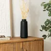 A black ceramic vase with dried flowers sits on a wooden cabinet next to a framed artwork and a book. Anteprima dell’immagine prodotto