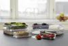 Several clear plastic food storage containers filled with food items like lettuce, crackers, grapes, cheese, and strawberries are arranged on a dark countertop in front of a window. Produktbild-Vorschau