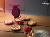 A mini waffle maker with various waffle plates and freshly made waffles are arranged on a wooden table, with raspberries and eggs in the foreground. Produktbild-Vorschau
