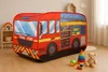 A children's play tent shaped like a fire truck is set up in a room with toys. The tent is red with yellow and blue accents, depicting windows, doors, and equipment. Toys like a stacking ring, a ball, and a teddy bear are placed around the tent. Produktbild-Vorschau