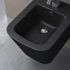 A close-up, angled view of a matte black bidet mounted on a wall, with a concrete wall and floor visible. Produktbild-Vorschau