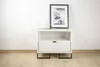 A white nightstand with a drawer and an open shelf, decorated with a framed picture and a cup on top, placed against a white wall. Podgląd zdjęcia produktu