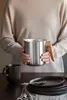 A person holding a stainless steel teapot with a wooden handle, with a dark mug and plates on a table. Náhled obrázku produktu