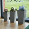 Three planters with flowers are placed on a wooden floor next to a large window with a view of a grassy area and a bicycle in the background. Produktbild-Vorschau