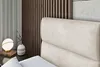 A close-up view of a bed with a beige upholstered headboard, a bedside table with a lamp and a vase, and a wooden slatted wall in the background. Produktbild-Vorschau