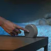 A hand is pressing a button on a small, round, gray device placed on a wooden table. The background is dark and appears to be a blurred natural setting with rocks and water. Voir les images du produit