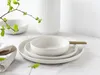 A minimalist still life of white ceramic tableware, including plates and bowls, set on a white tablecloth. A sprig of green leaves adds a touch of nature to the scene. Produktbild-Vorschau