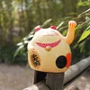 A beige Maneki-neko (lucky cat) figurine sits on a wooden railing, with blurred green foliage in the background. Produktbild-Vorschau