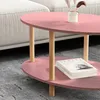 A pink oval coffee table with wooden legs is shown in a living room setting, next to a sofa. Books are placed on the lower shelf of the table. Podgląd zdjęcia produktu