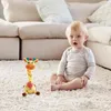Gelbes Plüsch-Giraffenspielzeug mit orangefarbener Schnauze und bunten Hörnern, sitzend auf einem weißen Teppich neben einem Baby Podgląd zdjęcia produktu