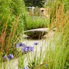 A garden scene with a reflective bowl as a water feature, surrounded by lush greenery and flowers. Produktbild-Vorschau
