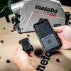 Hands holding a Metabo tracker and a smartphone displaying the Metabo Tracker app, with a Metabo power tool in the background. Voir les images du produit