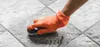 A gloved hand is scrubbing a tiled floor with a brush and soap. Náhled obrázku produktu
