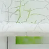 A close-up view of a white roman blind with a green branch pattern, partially covering a green window. Produktbild-Vorschau