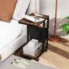 A C-shaped side table with storage is placed next to a bed, with items like magazines, boxes, and slippers around it. Voir les images du produit