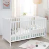 A white wooden baby crib with bedding and a grey blanket inside, placed on a rug in a nursery setting. Produktbild-Vorschau