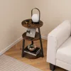 A three-tiered round side table with a rustic wood finish and black metal legs is placed in a corner next to a white sofa. The table is decorated with a lamp, a mug, a framed photo, and books. Podgląd zdjęcia produktu