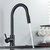 A black kitchen faucet with a pull-down sprayer is shown in use, with water flowing into a sink. Cups are hanging on a rack in the background. Produktbild-Vorschau