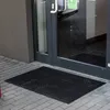 A black rubber doormat is placed on a tiled walkway in front of a glass door. Podgląd zdjęcia produktu