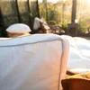 A close-up of a white pillow in a sunlit greenhouse with other pillows and furniture in the background. Produktbild-Vorschau