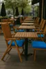 An outdoor seating area with wooden tables and chairs, some with blue cushions. Produktbild-Vorschau