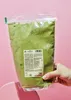 A hand holds a clear plastic bag filled with green powder against a pink background. Produktbild-Vorschau