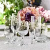 A collection of clear glass drinkware is arranged on a textured white surface, with a blurred floral arrangement in the background. Anteprima dell’immagine prodotto