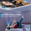 A woman is lying on a bed in a bedroom, looking at a projector that is projecting an image onto the ceiling. The projector is small and white. Produktbild-Vorschau