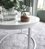 A close-up shot of a white round coffee table with a glass vase of greenery and a wooden decorative box on its surface, set on a textured gray rug. Náhľad obrázku produktu