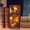 A miniature DIY book nook kit is displayed on a wooden surface next to several large, old books. The book nook is illuminated, showing a detailed interior with stairs, furniture, and cats. Voir les images du produit