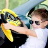 Gelb-schwarzes Spielzeuglenkrad mit Tachometer und Tasten, gehalten von einem Kind mit Sonnenbrille im Auto Voir les images du produit