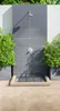 An outdoor shower area with a tiled wall, shower head, and a grate for drainage. Produktbild-Vorschau