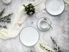 A flat lay of a white porcelain coffee service set with black trim, arranged on a textured gray surface with a cream-colored fabric runner and some greenery. Náhľad obrázku produktu