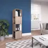 A tall, narrow, light brown wooden shelving unit stands against a vibrant blue wall in a room with a large window and a sofa. The shelves are organized with binders and books. A large potted plant is to the left of the unit, and a geometric rug is on the f Náhled obrázku produktu