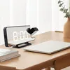 A digital alarm clock with a projector is placed on a wooden desk next to a laptop and books. Náhled obrázku produktu