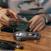A person's hands are shown working on electronic components on a wooden table, with a digital microscope in the foreground. Anteprima dell’immagine prodotto