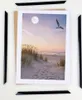 Four black frame pieces are arranged around a picture of a beach scene at sunset, with a seagull flying overhead. Produktbild-Vorschau