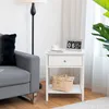 A white nightstand with a drawer and an open shelf is placed next to a gray sofa in a living room setting. Podgląd zdjęcia produktu