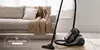 A vacuum cleaner is placed on a rug in front of a sofa in a living room. Podgląd zdjęcia produktu
