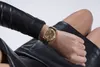 A close-up shot of a woman's wrist wearing a gold Guess watch, with her legs in black leather clothing visible. Náhled obrázku produktu
