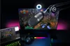 A gaming setup with a microphone in the foreground, a laptop and monitor displaying games in the background, and colorful LED lighting. Podgląd zdjęcia produktu