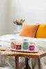 Four candles are displayed on a wooden bench in front of a bed with pillows. Produktbild-Vorschau