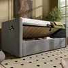 A grey upholstered sofa bed with its storage compartment open, revealing stored items. A phone is charging via a USB cable plugged into a side pocket. The bed is in a room with a patterned rug and a window in the background. Anteprima dell’immagine prodotto
