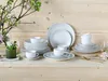 A collection of white porcelain dinnerware, including plates, bowls, and cups, arranged on a wooden surface with a floral arrangement in the background. Produktbild-Vorschau