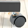 A close-up of a black caster wheel attached to a black metal frame, resting on a light wood-grain floor. Orange arrows indicate rotation. Náhled obrázku produktu